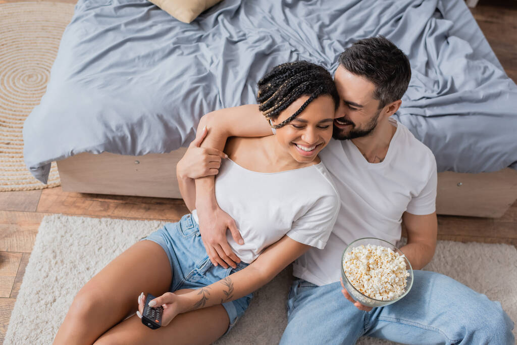 high angle view of cheerful man with bowl of popcorn embracing african american woman with tv remote controller on floor in bedroom - Photo, Image