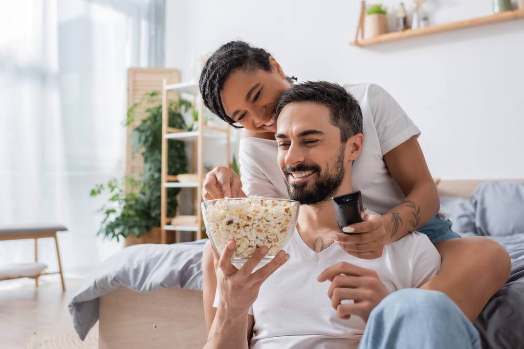 heureuse femme afro-américaine avec télécommande tv prendre popcorn près de sourire homme barbu dans la chambre à coucher à la maison - Photo, image