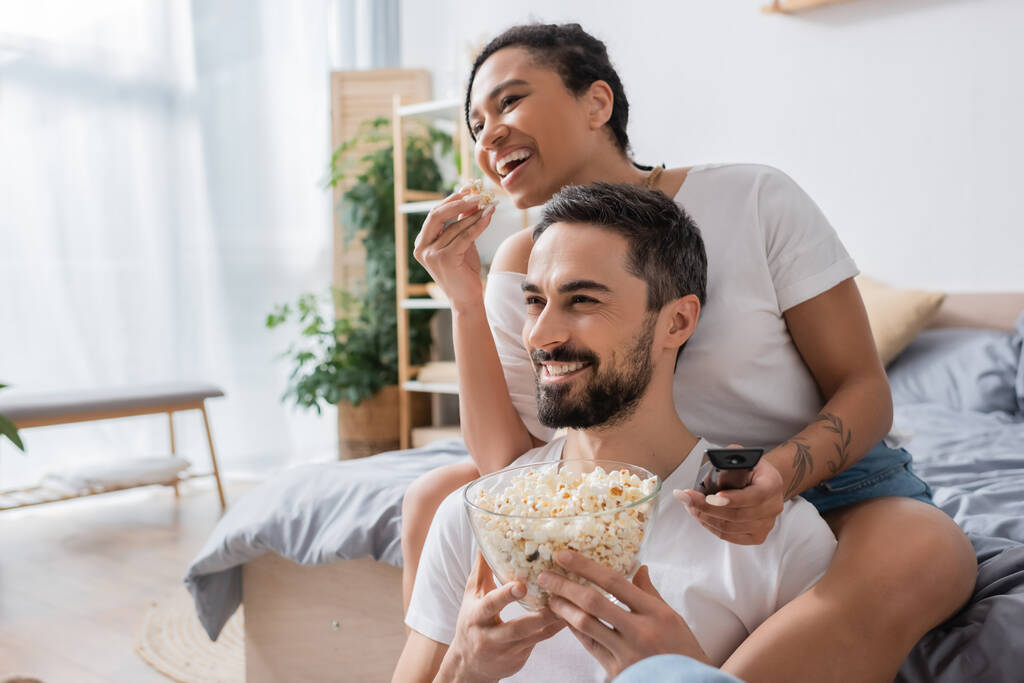 pareja interracial complacido comer palomitas de maíz y ver películas en el dormitorio en casa - Foto, Imagen