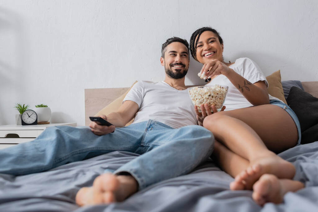 задоволена афроамериканська жінка їсть попкорн біля усміхненого бородатого чоловіка з телевізійним пультом дистанційного керування на ліжку вдома
 - Фото, зображення