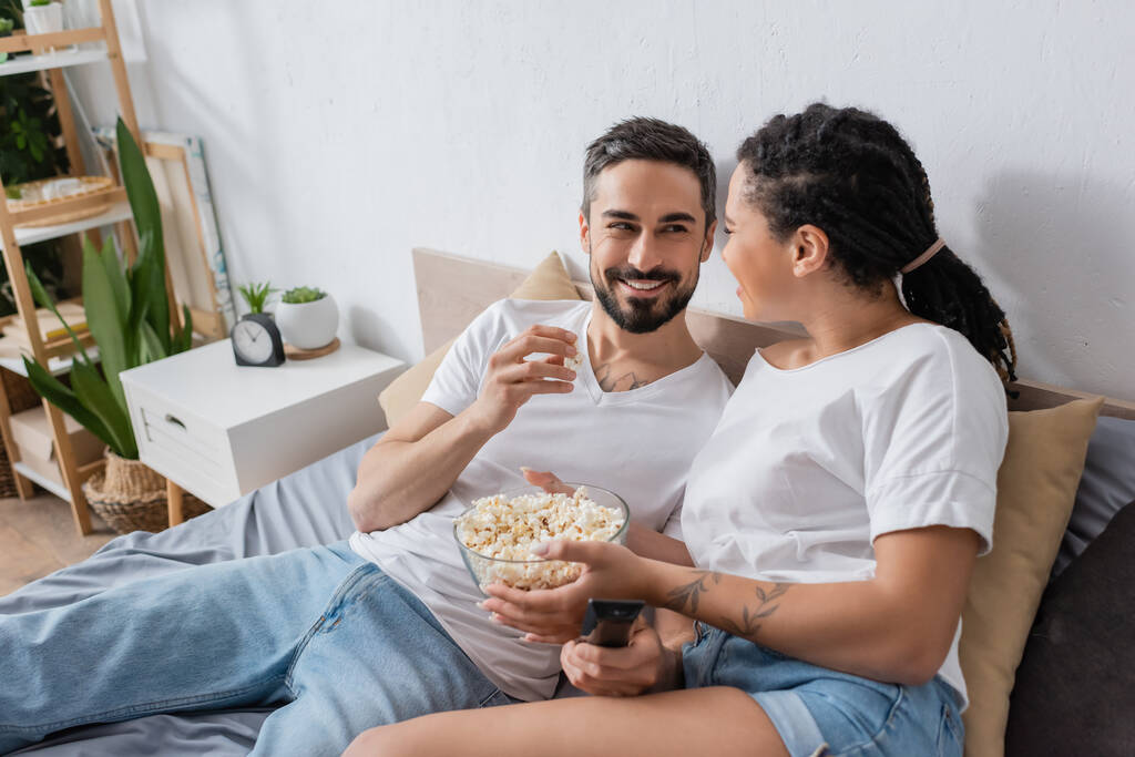 allegra coppia interrazziale con telecomando tv e popcorn sorridenti a vicenda sul letto a casa - Foto, immagini