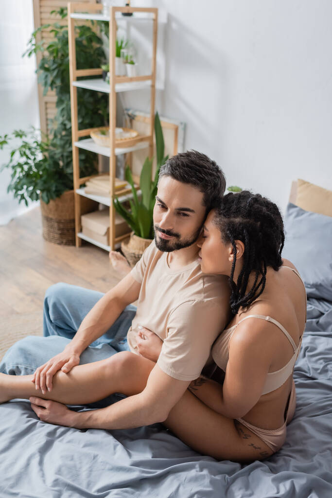 sexy african american woman in lingerie seducing bearded man sitting in t-shirt and jeans on bed at home - Photo, Image