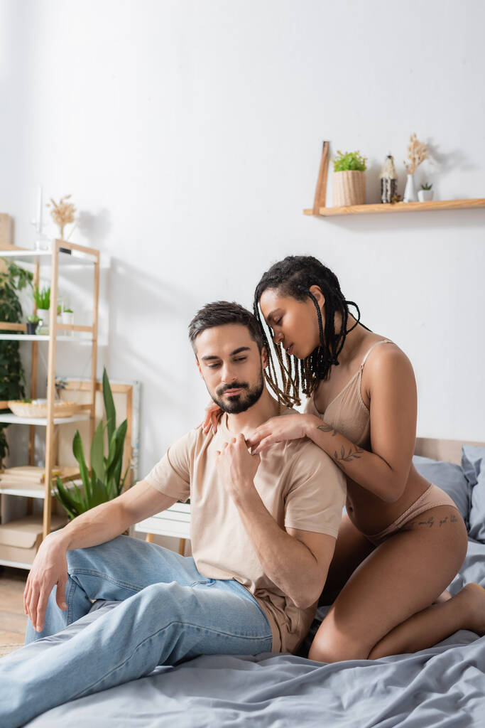 szakállas férfi pólóban és farmerban, aki egy tetovált afro-amerikai nő kezét fogja fehérneműben otthon a hálószobában. - Fotó, kép