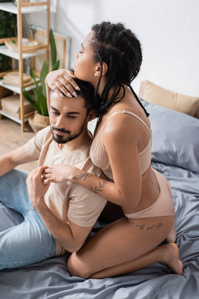 femme afro-américaine tatouée en lingerie embrassant petit ami barbu assis en t-shirt et jeans sur le lit à la maison - Photo, image