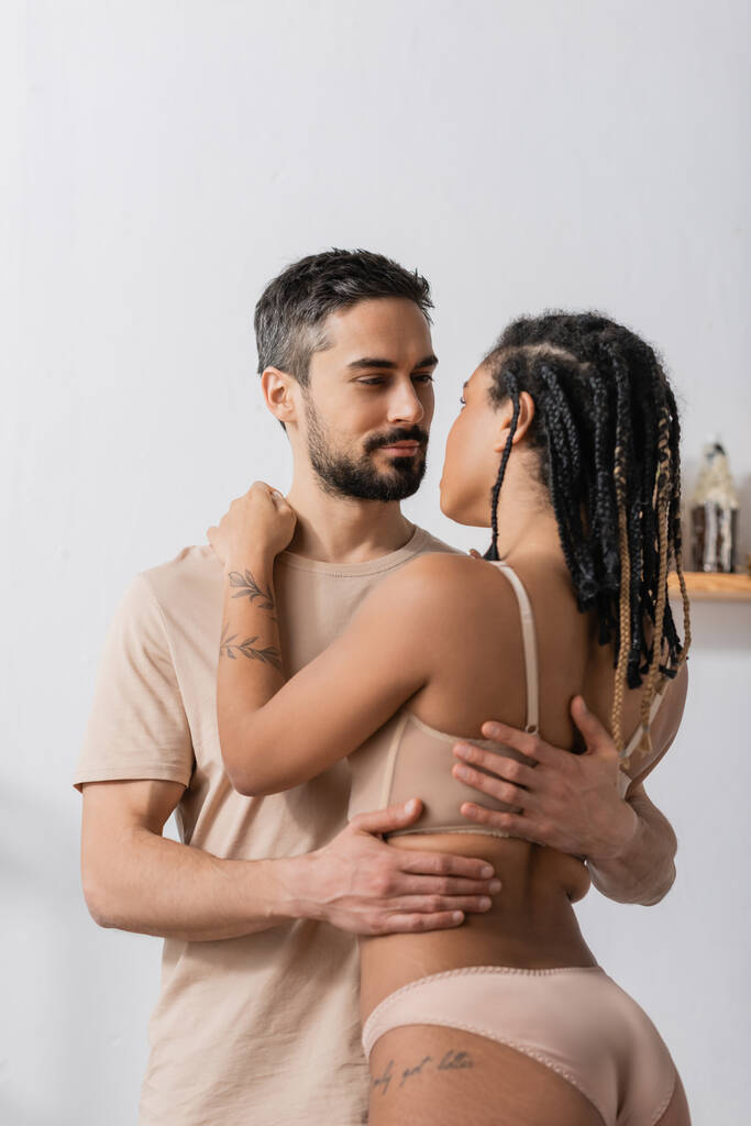 бородатий чоловік у футболці обіймає спокусливу афроамериканську жінку в нижній білизні в спальні вдома
 - Фото, зображення
