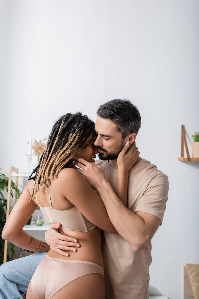 leidenschaftliche afrikanisch-amerikanische Frau in Dessous küsst mit bärtigen Mann im T-Shirt im Schlafzimmer zu Hause - Foto, Bild