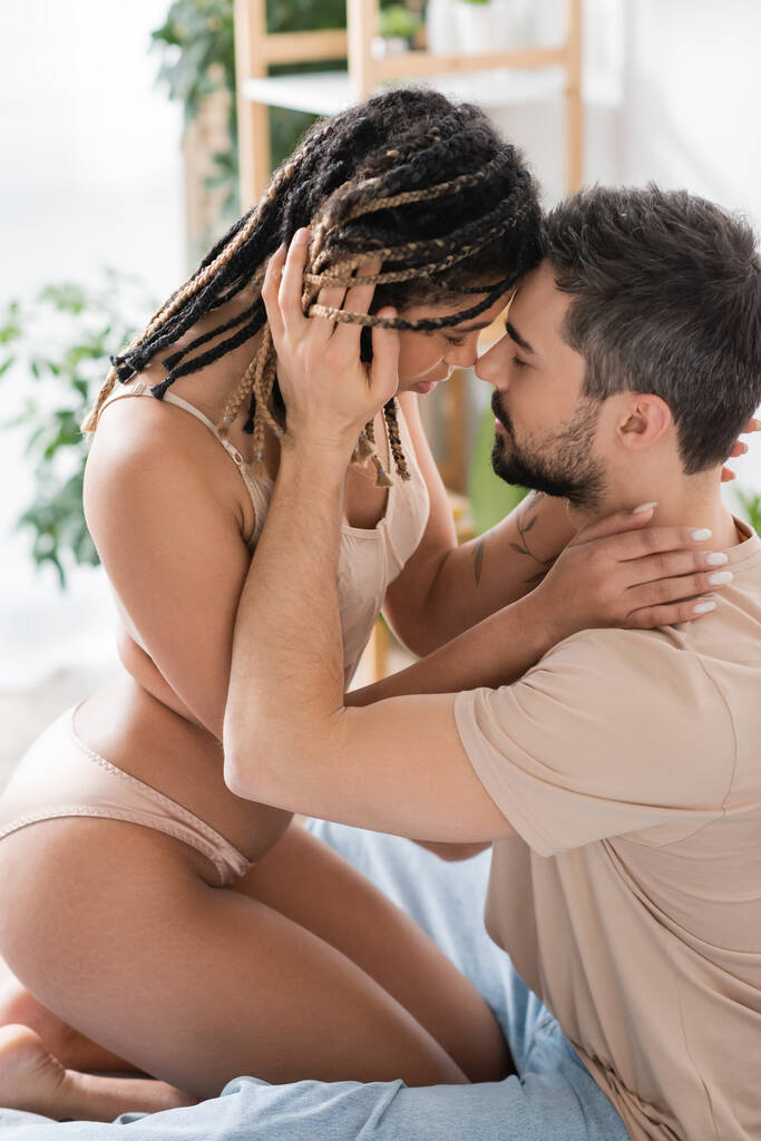boční pohled na vášnivé africké Američanky v sexy spodní prádlo v blízkosti brunetky muž v tričku v ložnici doma - Fotografie, Obrázek
