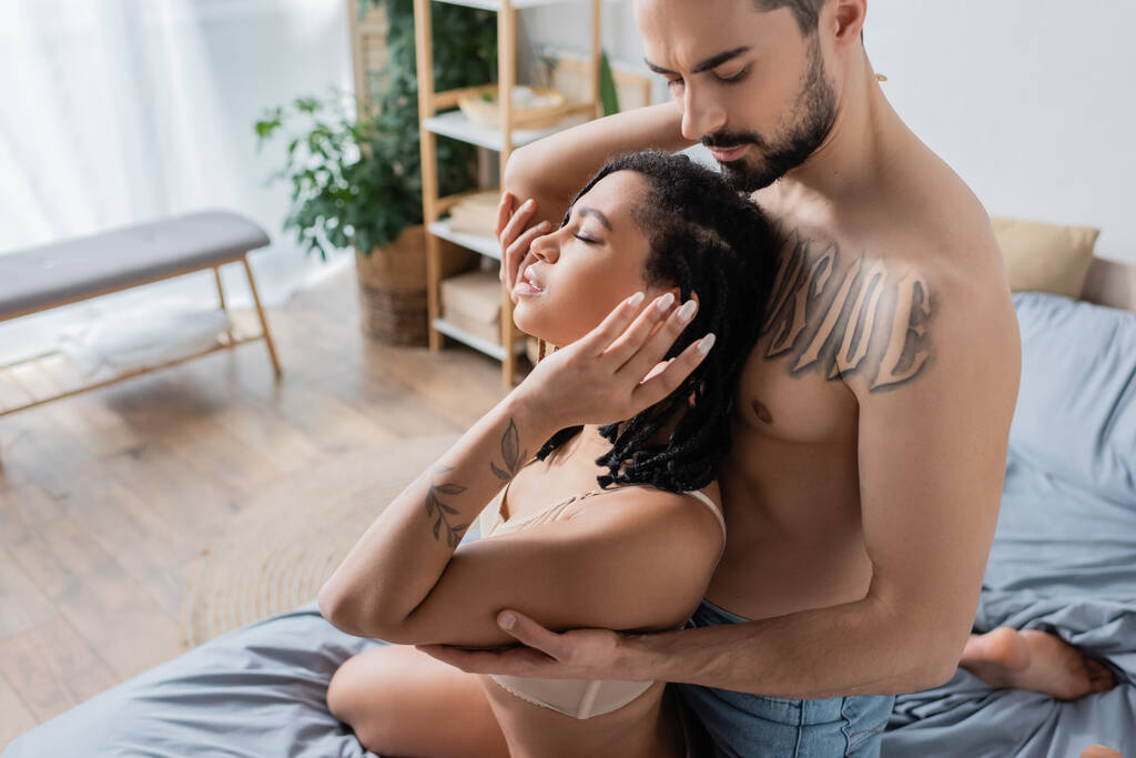 puolella näkymä paidaton tatuoitu mies käsittää seksikäs afrikkalainen amerikkalainen nainen alusvaatteet makuuhuoneessa kotona - Valokuva, kuva