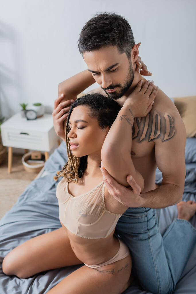 νέα και καυτή Αφροαμερικανή γυναίκα με σουτιέν αγκαλιάζει γενειοφόρο γυμνό άντρα στο υπνοδωμάτιο στο σπίτι - Φωτογραφία, εικόνα