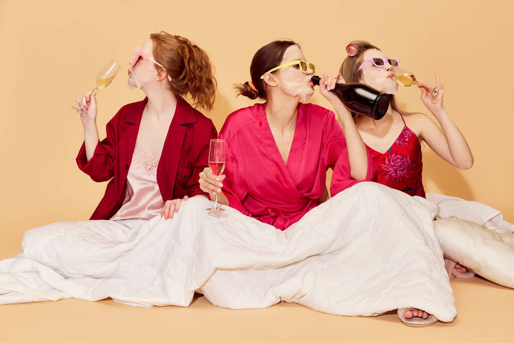 Chicas jóvenes elegantes y bonitas en pijama con máscaras faciales, sentadas debajo de una manta en la pijamada y bebiendo champán. Concepto de juventud, emociones, belleza, amistad, fiesta, relajación, sentimientos - Foto, Imagen