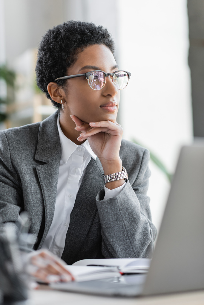 pensativa mulher de negócios afro-americana em blazer cinza e óculos olhando para longe no escritório - Foto, Imagem