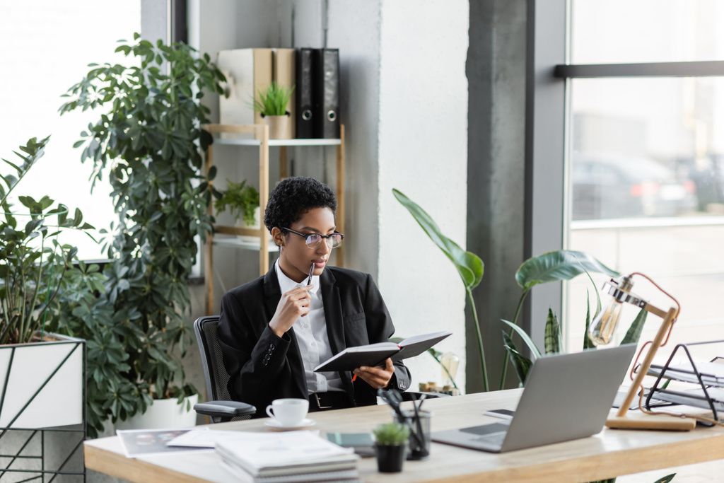 przemyślana afrykańska bizneswoman trzyma pióro i patrząc na notatnik w pobliżu laptopa i dokumentów w nowoczesnym biurze - Zdjęcie, obraz