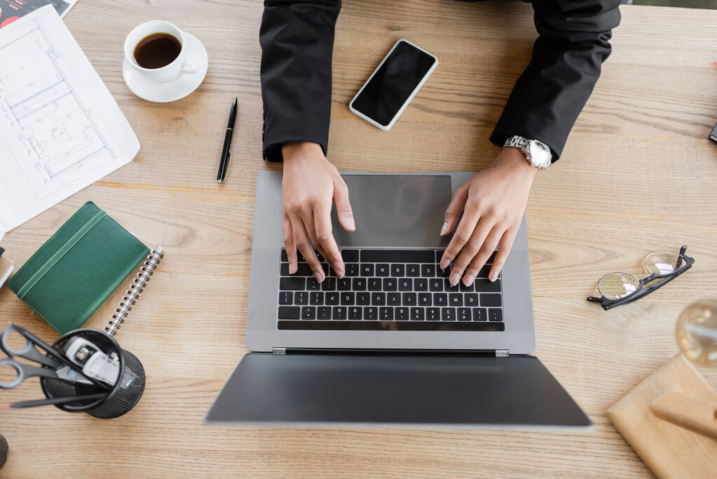 vrchní pohled na oříznutý africký americký manažer psaní na notebooku v blízkosti smartphone a šálek kávy na pracovním stole v kanceláři - Fotografie, Obrázek