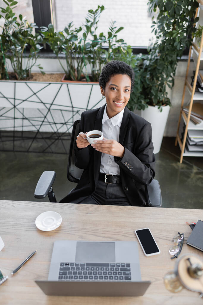 wysoki kąt widzenia udanej afrykańskiej bizneswoman uśmiecha się do kamery siedząc z filiżanką kawy w pobliżu gadżetów - Zdjęcie, obraz