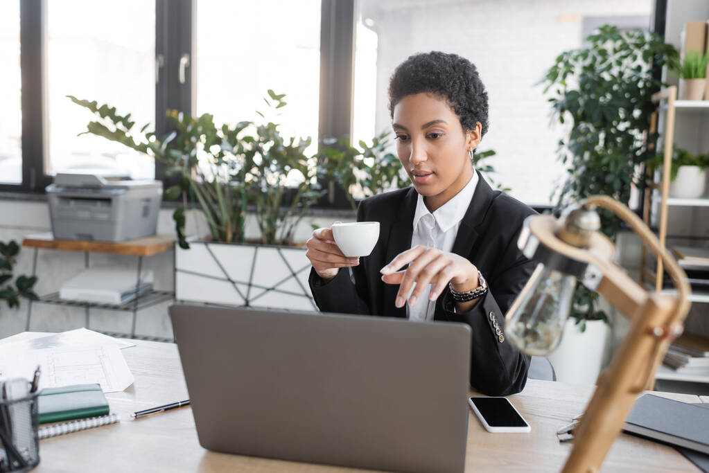 jeune femme d'affaires afro-américaine avec tasse de café pointant vers un ordinateur portable près du smartphone et lampe de bureau floue dans le bureau moderne - Photo, image