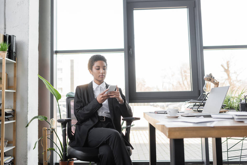 stylový africký americký podnikatelek zprávy na mobilním telefonu v blízkosti notebooku na pracovním stole v moderní kanceláři - Fotografie, Obrázek