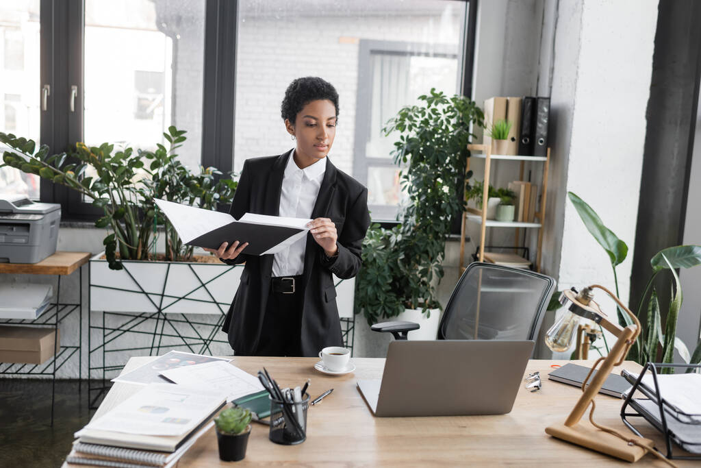 mulher de negócios americana africana em preto pasta terno segurando com documentos e olhando para laptop na mesa de trabalho no escritório moderno - Foto, Imagem