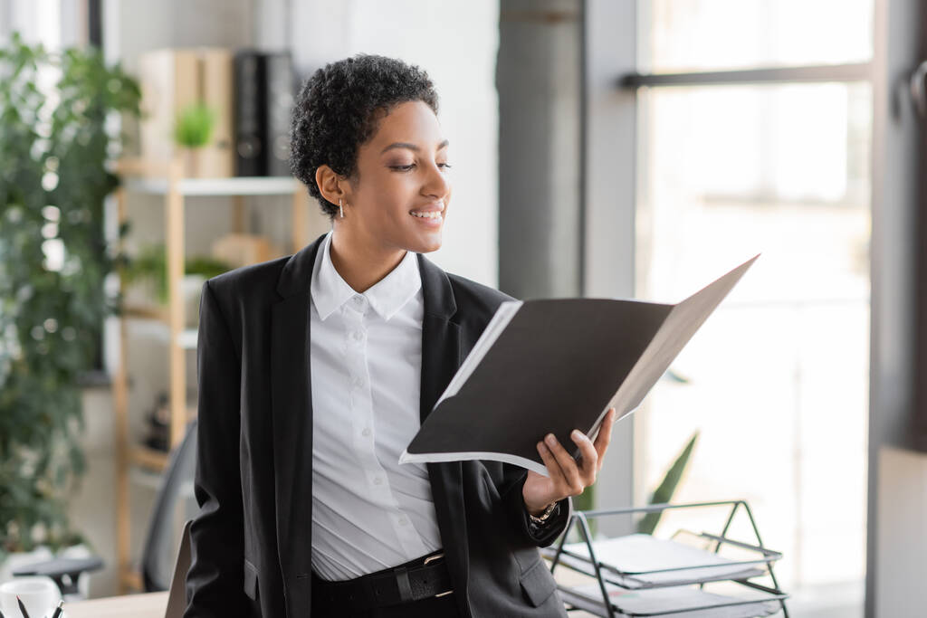 zadowolony afrykański amerykański bizneswoman w czarny blezer patrząc na dokumenty w folderze podczas stojąc w biurze - Zdjęcie, obraz