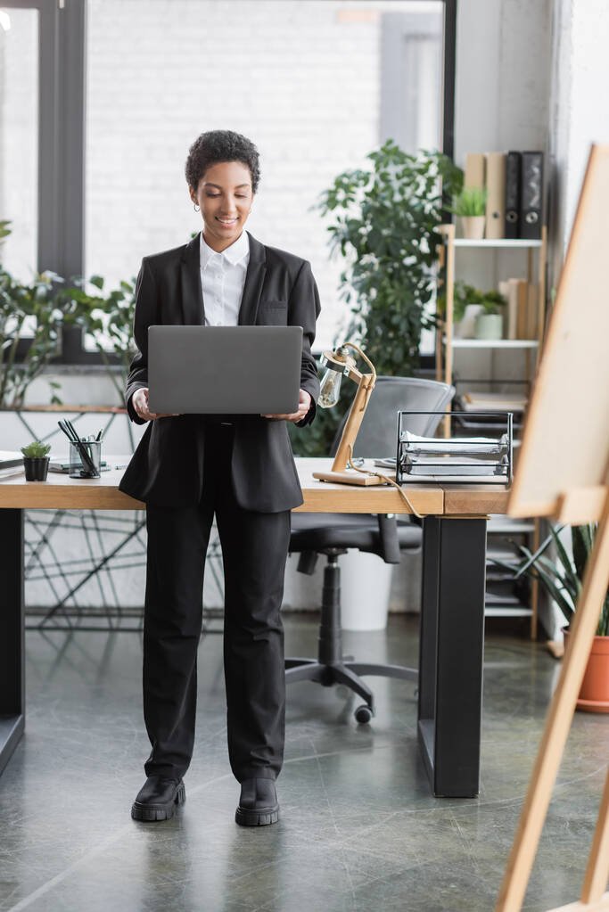 longitud completa de la exitosa mujer de negocios afroamericana en traje negro que sostiene el ordenador portátil mientras está de pie en el escritorio de trabajo en la oficina - Foto, imagen