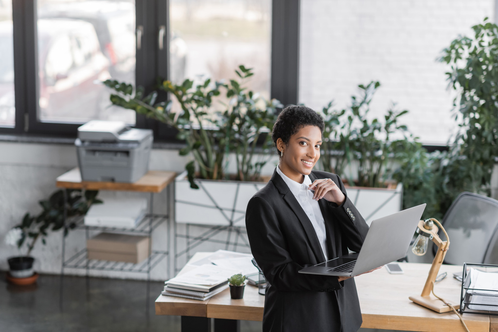 vreugdevolle Afrikaanse Amerikaanse zakenvrouw op zoek naar camera terwijl staande met laptop in de buurt van het werk bureau in modern kantoor - Foto, afbeelding