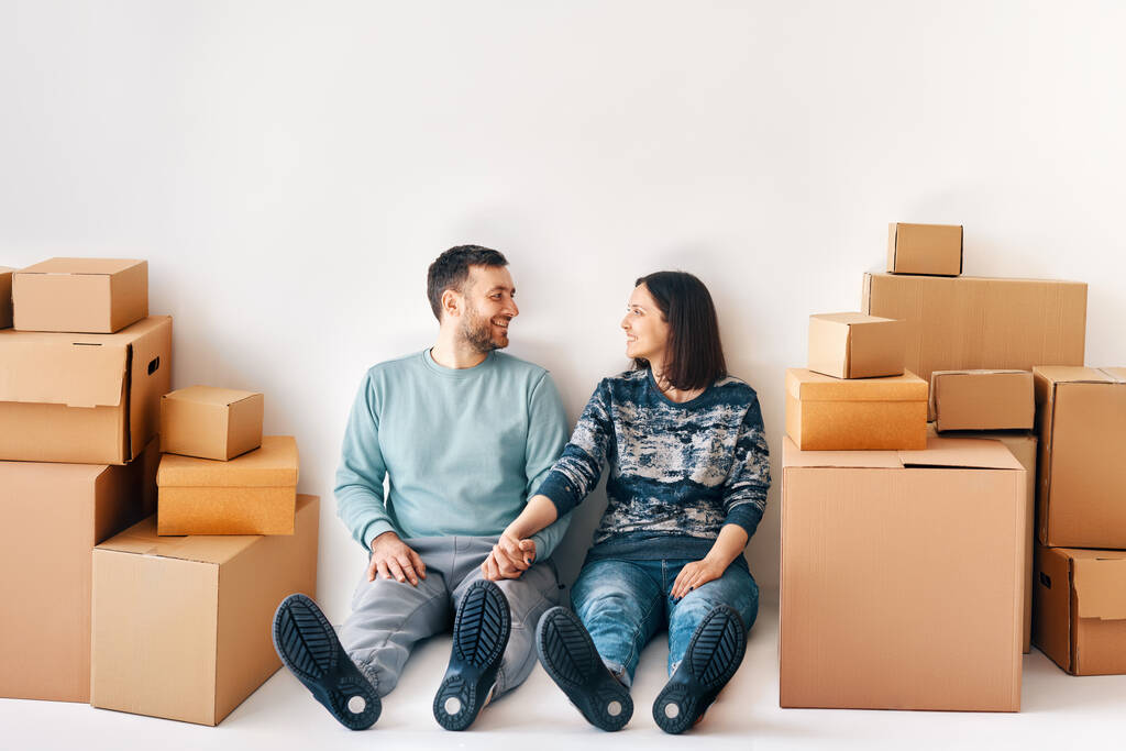 Happy smiling couple at new home sitting on floor surrounded by cardboard boxes. Moving day and interior renovation concept - Photo, Image
