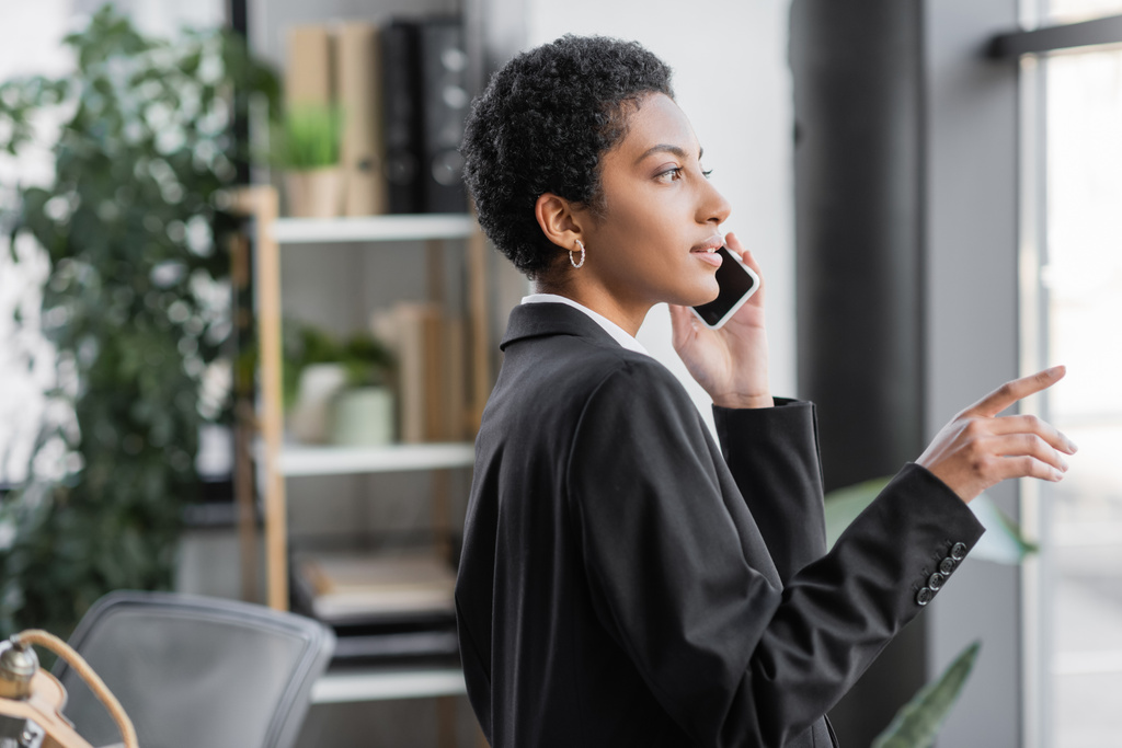 side view of young african american american businesswoman in black blazer pointing with finger and talking on cellphone in office - Photo, Image