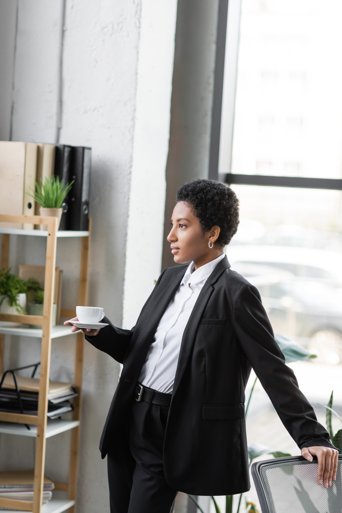młody afrykański amerykański bizneswoman w czarny garnitur stojąc z filiżanką kawy i patrząc w górę w biurze - Zdjęcie, obraz