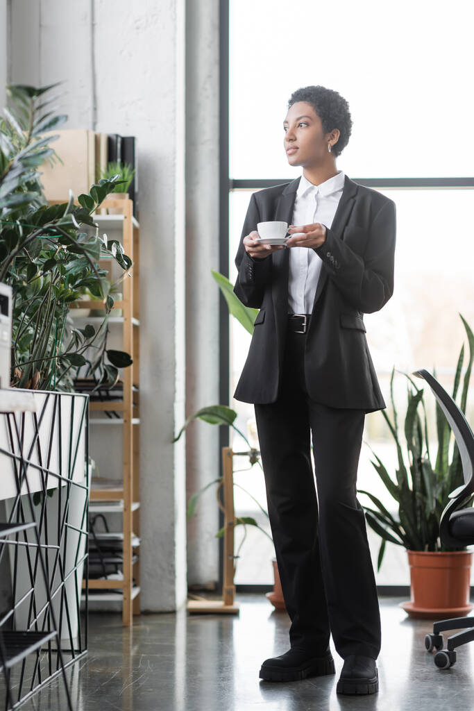 longitud completa de la mujer de negocios afroamericana en traje negro de pie con taza de café cerca de plantas en maceta verde en la oficina moderna - Foto, Imagen