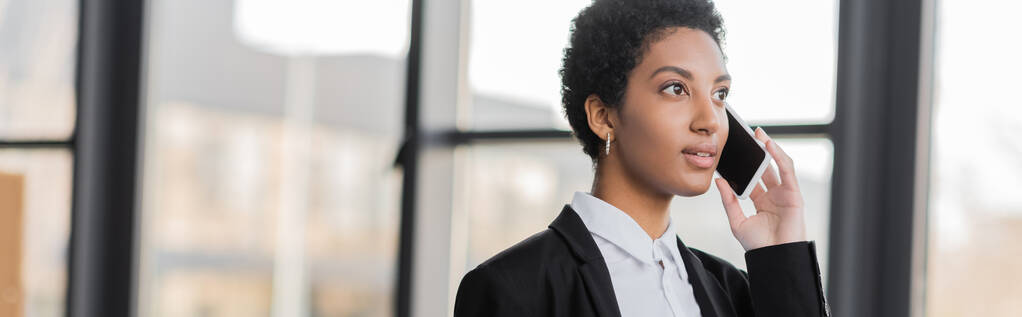mladá africká americká podnikatelka odvrací pohled, zatímco mluví na mobilním telefonu v kanceláři, banner - Fotografie, Obrázek