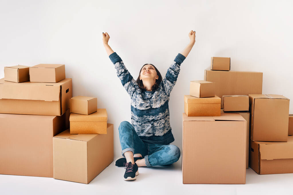 La joven feliz celebra mudarse a un nuevo hogar con las manos levantadas sentadas en el suelo rodeadas de cajas de cartón. Bienes raíces, concepto de reubicación - Foto, imagen