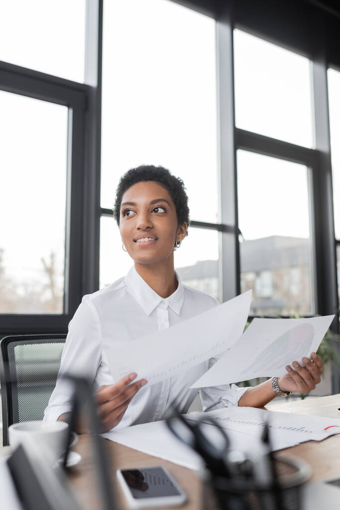 χαρούμενη αφροαμερικανίδα επιχειρηματίας με λευκή μπλούζα που κρατάει έγγραφα και κοιτάζει αλλού στο γραφείο - Φωτογραφία, εικόνα