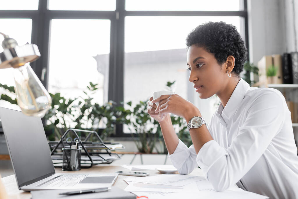 vista lateral da empresária americana africana segurando xícara de café e olhando para laptop com tela em branco no escritório - Foto, Imagem