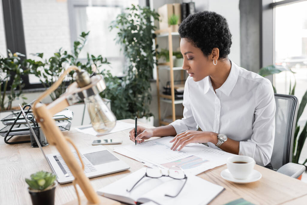 νεαρή αφροαμερικανίδα επιχειρηματίας με λευκή μπλούζα που γράφει σε έγγραφα κοντά σε laptop και καφέ κύπελλο στο σύγχρονο γραφείο - Φωτογραφία, εικόνα