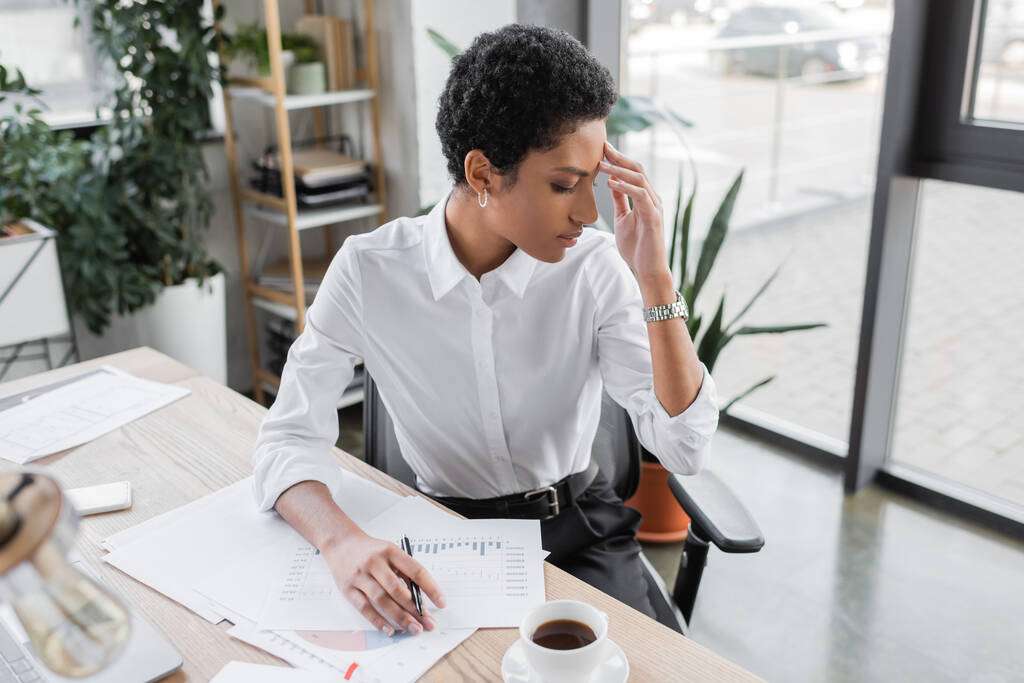 κουρασμένη αφροαμερικανίδα επιχειρηματίας κάθεται κοντά σε έγγραφα με διαγράμματα και πάσχουν από πονοκέφαλο στο γραφείο - Φωτογραφία, εικόνα