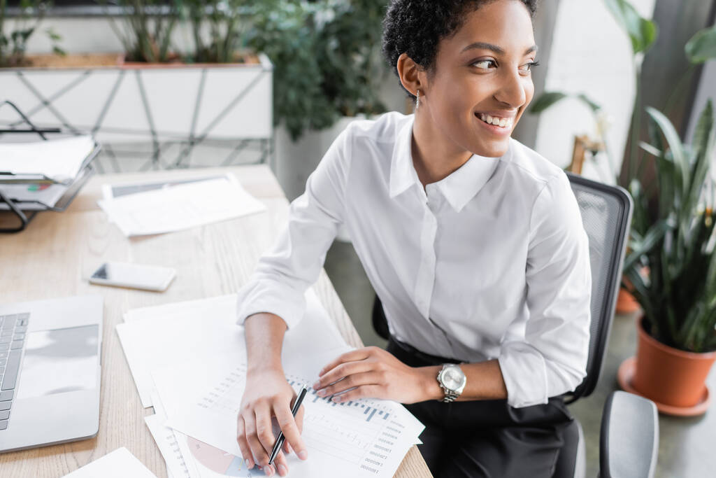 Весела афроамериканська комерсантка сидить біля документів і технічних приладів і озирається в офіс - Фото, зображення