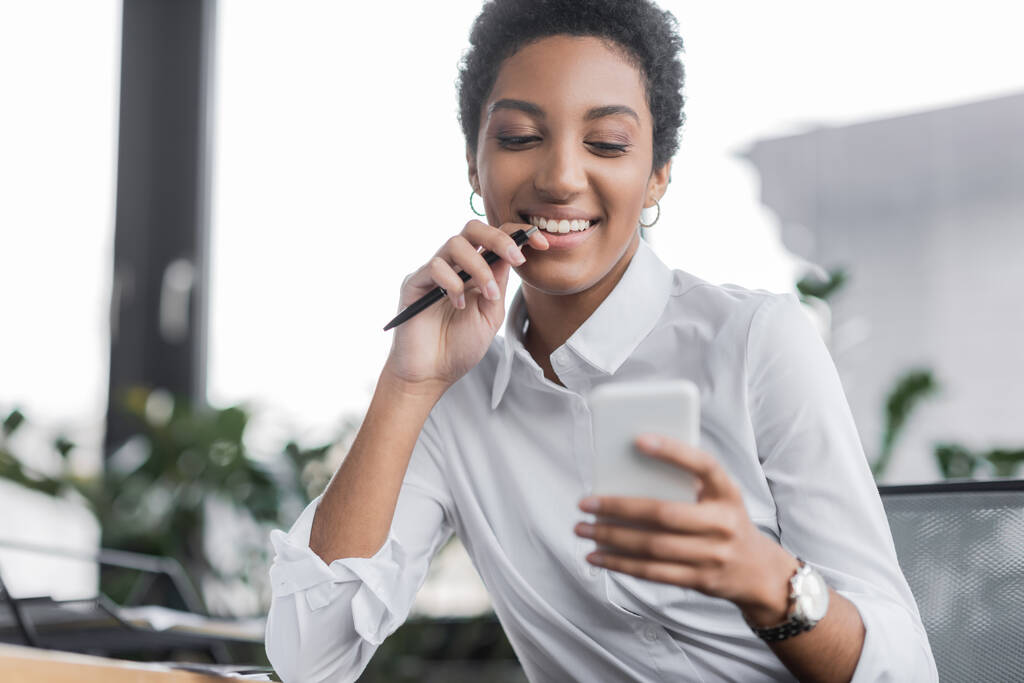 alegre mujer de negocios afroamericana en blusa blanca sosteniendo pluma y mirando el teléfono móvil en la oficina - Foto, Imagen