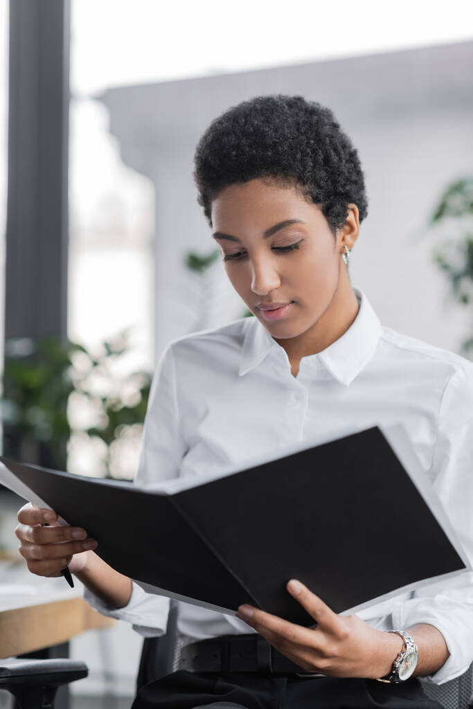 jeune femme d'affaires afro-américaine en chemisier blanc lisant des documents dans le dossier au bureau - Photo, image