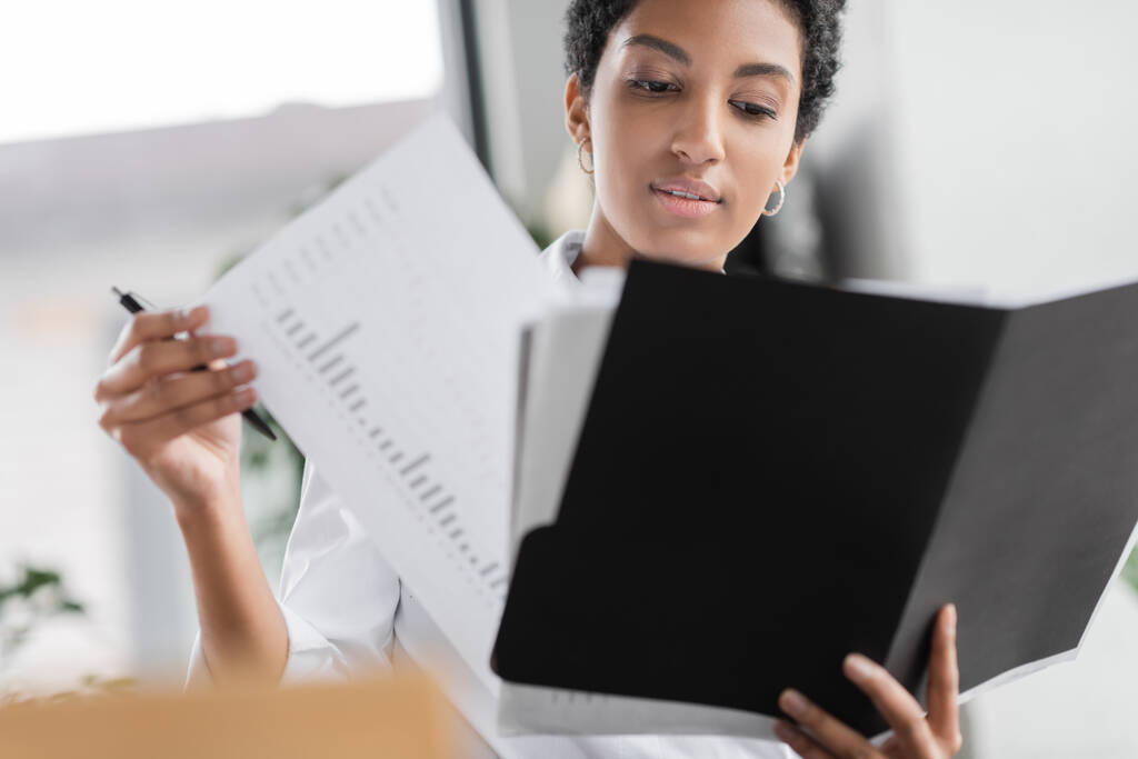 νεαρή αφροαμερικανίδα επιχειρηματίας κρατώντας φάκελο και κοιτάζοντας τα χαρτιά με infographics στο γραφείο - Φωτογραφία, εικόνα