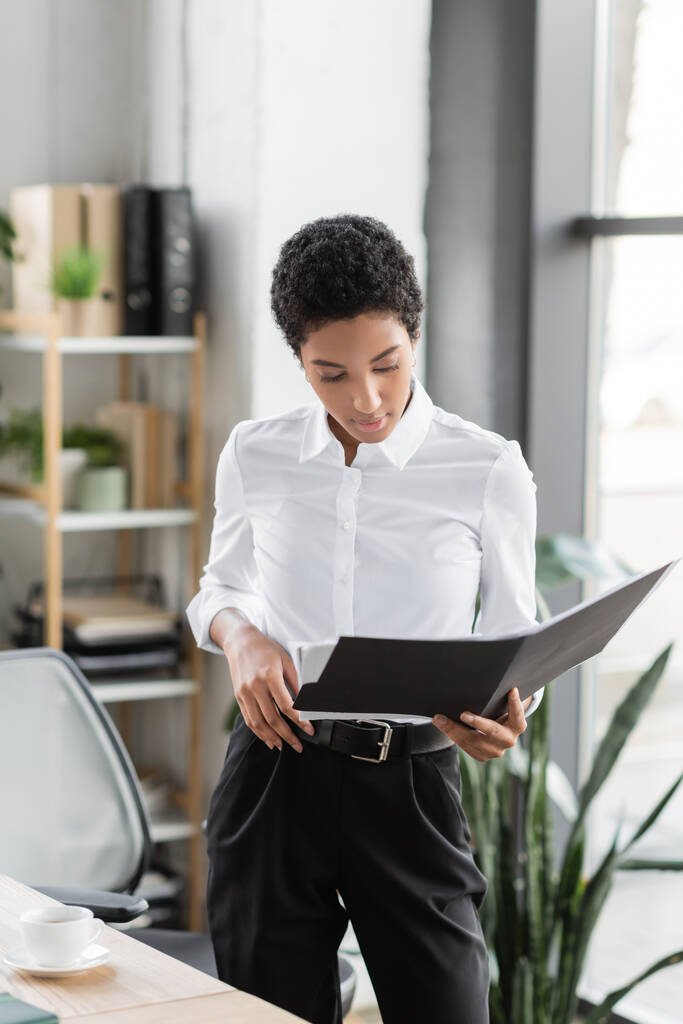 elegante donna d'affari africana americana in camicetta bianca e pantaloni neri in piedi con cartella in ufficio - Foto, immagini