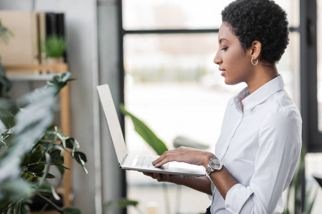 Genç Afro-Amerikan iş kadınının ofiste dikilip boş ekranlı dizüstü bilgisayar kullanışının yan görüntüsü - Fotoğraf, Görsel