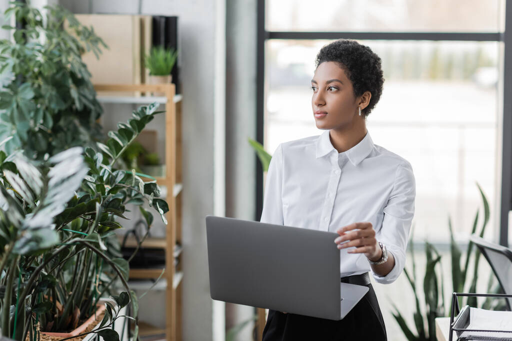 pensive african american businesswoman with laptop looking away while standing near green plants in modern office - Photo, Image