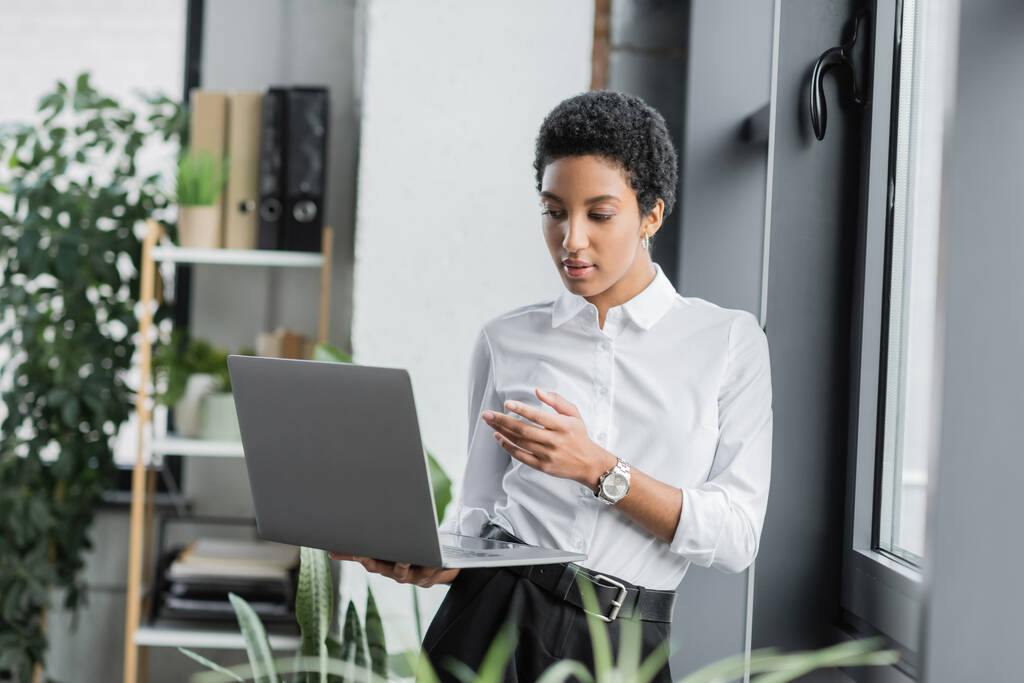 юна афроамериканська бізнесменка у білій блузці, яка показує рукою під час відеобесіди на ноутбуці при офісі. - Фото, зображення