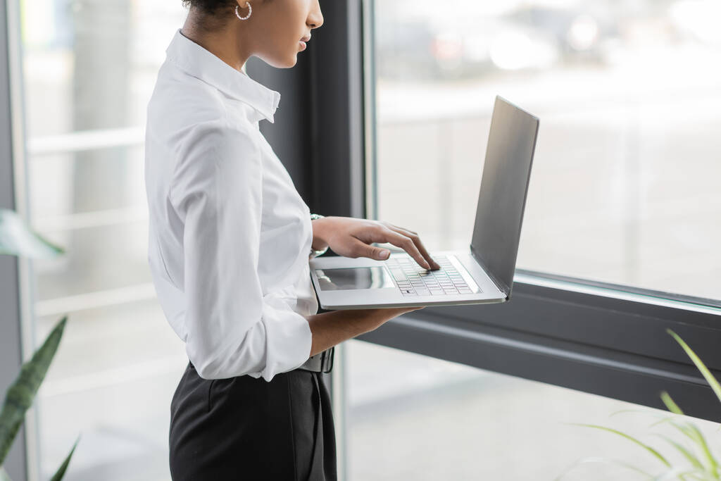 cropped άποψη της Αφρικής αμερικανική επιχειρηματίας σε λευκή μπλούζα με τη χρήση φορητού υπολογιστή με λευκή οθόνη κοντά στο παράθυρο στο γραφείο - Φωτογραφία, εικόνα