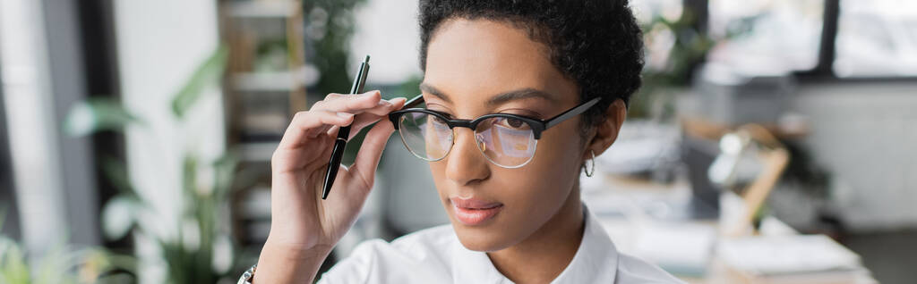 jovem mulher de negócios afro-americana segurando caneta e ajustando óculos elegantes no escritório, bandeira - Foto, Imagem