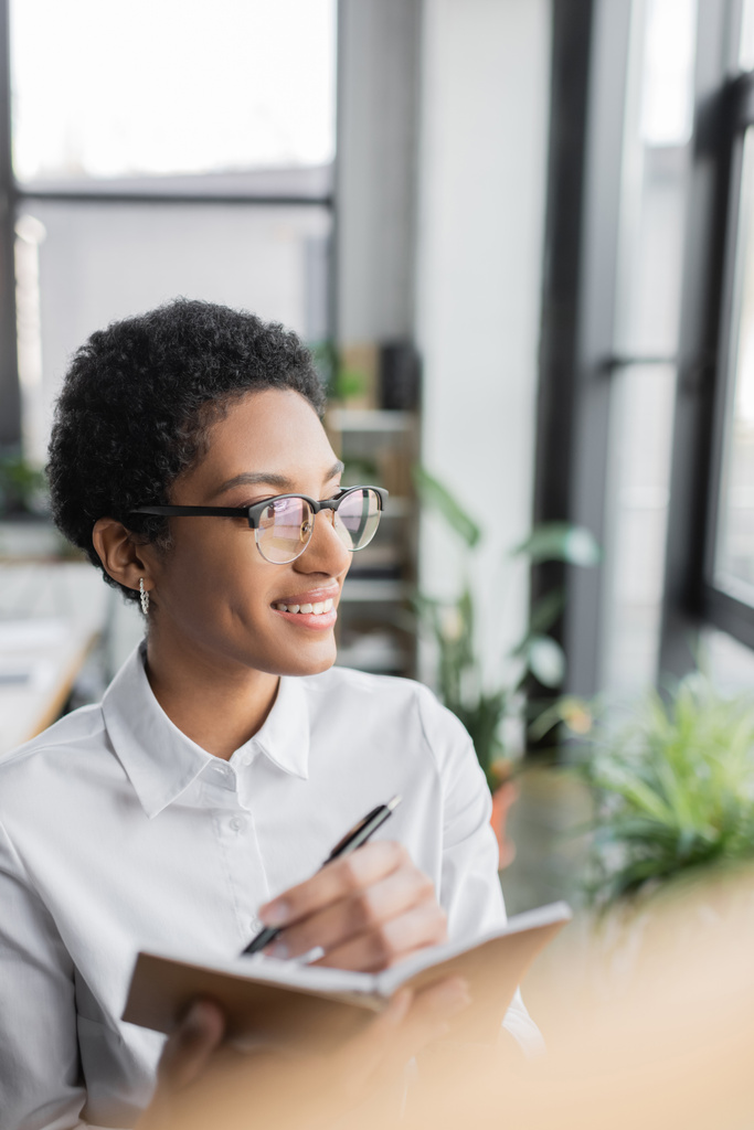 mulher de negócios americana africana alegre em óculos elegantes e blusa branca escrevendo em notebook e olhando para longe no escritório - Foto, Imagem