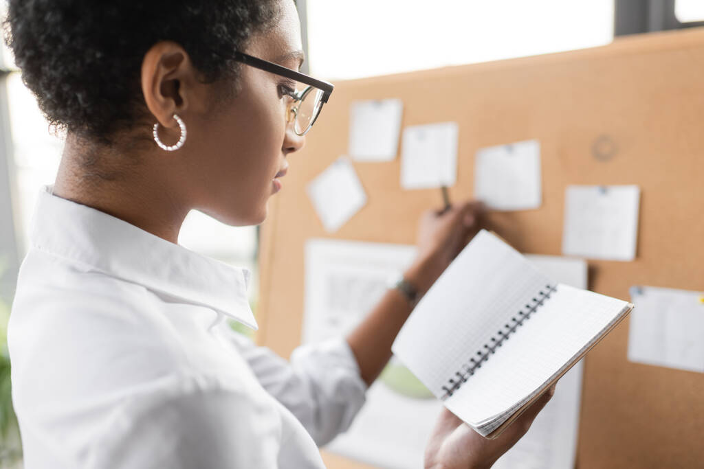 side view of african american businesswoman standing near blurred note board and holding blank notebook in office - Photo, Image