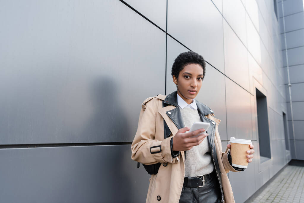 femme d'affaires afro-américaine à la mode avec tasse jetable et smartphone regardant la caméra près du mur gris sur la rue en ville - Photo, image