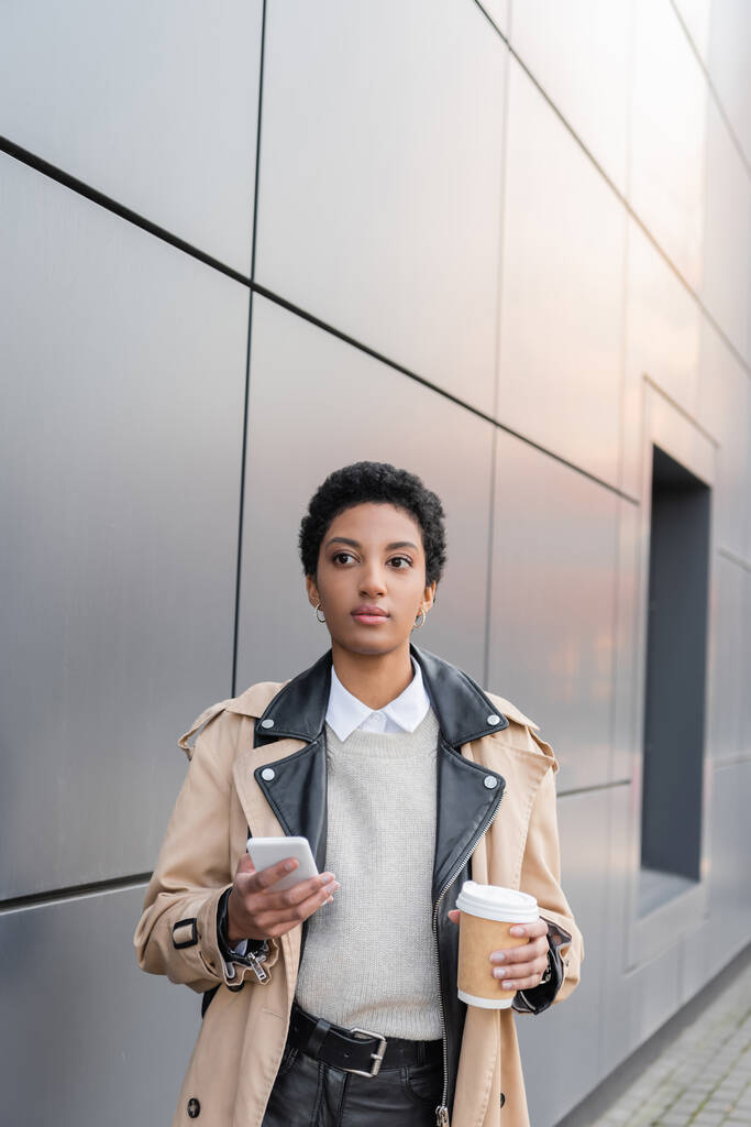 νεαρή αφροαμερικανίδα επιχειρηματίας στη μόδα καμπαρντίνα κρατώντας smartphone και καφέ για να πάει κοντά γκρίζο τοίχο σε αστικό δρόμο - Φωτογραφία, εικόνα