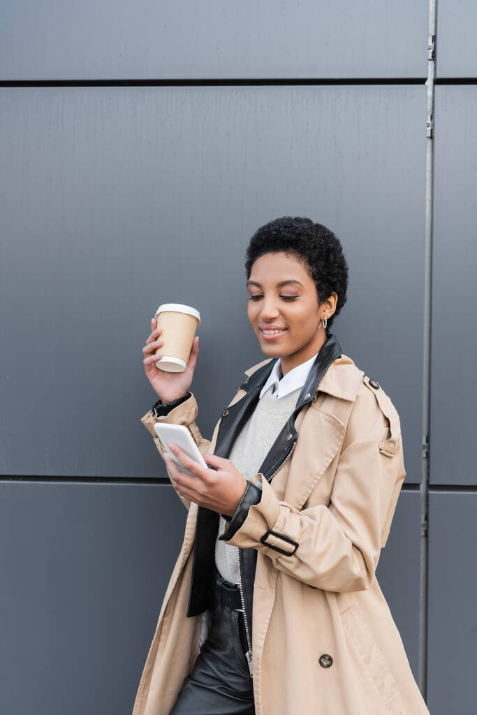 pozitív afro-amerikai üzletasszony bézs ballon kabát csevegés okostelefon és gazdaság elvitelre ital közelében szürke fal az utcán - Fotó, kép