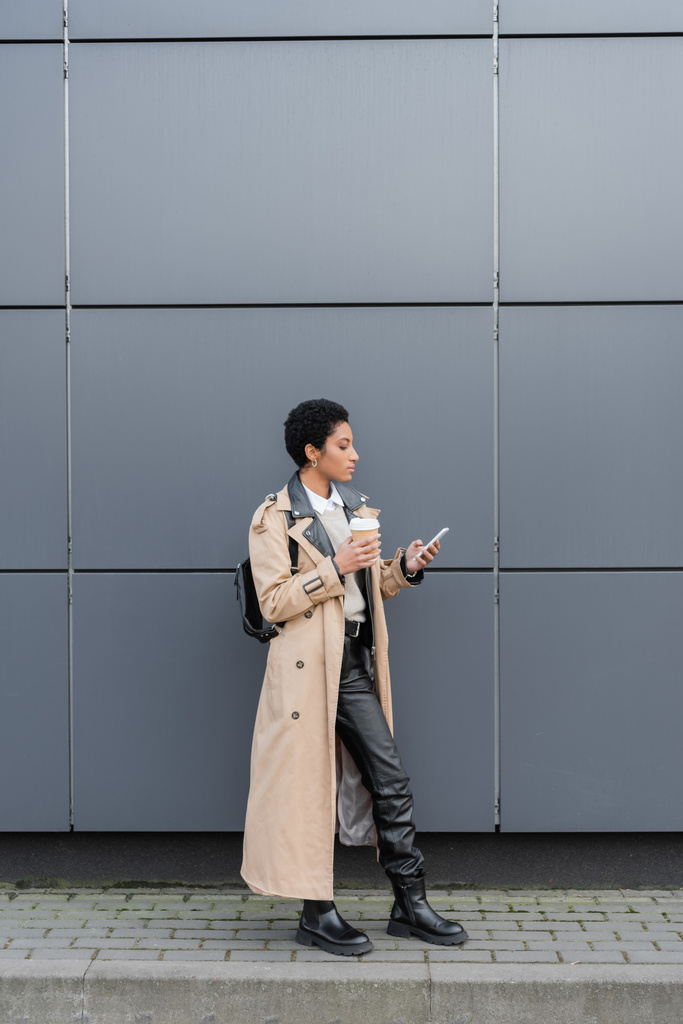 mujer de negocios afroamericana en gabardina y botas de cuero sosteniendo taza desechable y charlando en el teléfono celular en la calle - Foto, imagen